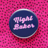 A round navy blue pinback button that reads NIGHT BAKER in a chunky pink script font, with a white drop shadow. Button is pinned to a vintage pink shirt with tiny black triangles