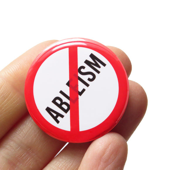 A round white button that reads ABLEISM in black text with a red circle and slash to indicate being against endless greed. Button badge is held in a hand.