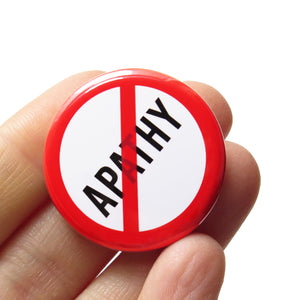 A round white button that reads No APATHY in black text with a red circle and slash to indicate being against endless greed. Button is held in a hand.