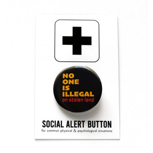 Round black pinback button that reads: NO ONE IS ILLEGAL ON STOLEN LAND in mustard yellow and red-orange text. Button is held in a hand.