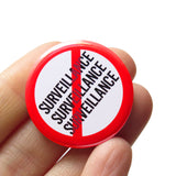 A round white button that reads NO SURVEILLANCE 3 times stacked,  in black text with a red circle and slash to indicate being against Surveillance. Badge is held in a hand.