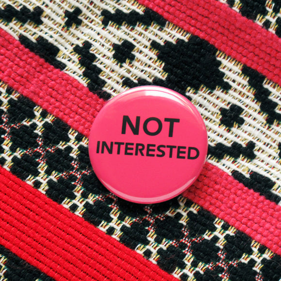 Round, dark pink magenta pinback button that reads NOT Interested in black text in 2 lines, NOT being larger. Button is on a black white and pink woven fabric,