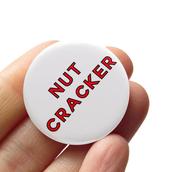 White round pinback button that reads NUTCRACKER in red san serif text.  Button is held in a hand