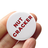 White round pinback button that reads NUTCRACKER in red san serif text.  Button is held in a hand