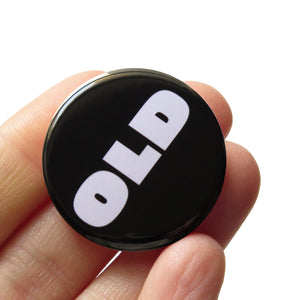 A round black pinback button that reads OLD in a chunky white font. Button is held in a hand.