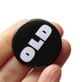 A round black pinback button that reads OLD in a chunky white font. Button is held in a hand.