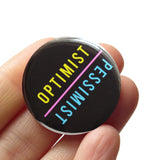 Round pinback buttons that says OPTIMIST, in yellow text, in one direction, and PESSIMIST, in blue text,  in the opposite direction.  Text is on a black background. The button is held in a hand.