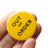 Round yellow 1.25" pinback button that reads OUT OF ORDER in black text. The button is held in a hand.