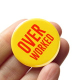 Round yellow pinback button that reads OVER WORKED in two lines of red text. button held in a hand.