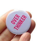  Round light gray button that reads OVER THINKINER in two lines in pink text. Badge is held in a hand.