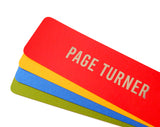 Four bright colored bookmarks fanned out, the top one is a bright red-orange bookmark that reads PAGE TURNER in gunmetal silver embossed text.And there is a marigold yellow, bright blue and deep moss green bookmarks fanned below, with no visible text.