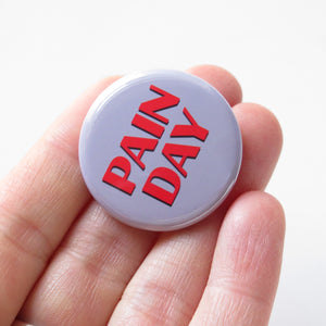 Round gray pinback button that reads PAIN DAY in a chunky red font with black drop shadow. Button is head in a hand by the fingers.