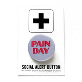 Round gray pinback button that reads PAIN DAY in a chunky red font with black drop shadow. Button is on a Social Alert Button Backing card.