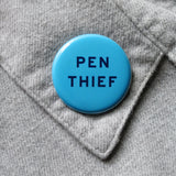 bright blue round pinback button that reads PEN THIEF in a dark blue san serif font. Button is pinned to a gray flannel shirt collar