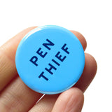 bright blue round pinback button that reads PEN THIEF in a dark blue san serif font. Button is held in a hand.