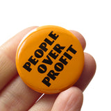 Round yellow-orange pinback button with black text that reads PEOPLE OVER PROFIT in an art deco font. Button is held in a hand.