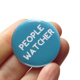 Round teal button that reads PEOPLE WATCHER in white text. Button is held in a hand.