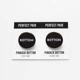 Two pinback buttons on a backing card with a center perforation to tear & share with a friend. Button the left reads BOTTOM. Button on right reads BOTTOM. Both buttons are black with white text.