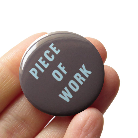 Dark gray button with mint green text reading PIECE OF WORK. Button is held in a hand.