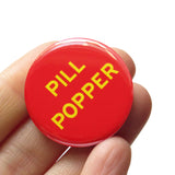 Round red 1.25" pinback button that reads PILL POPPER in yellow text. Button is held I a hand