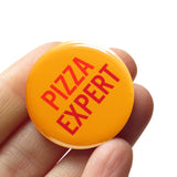Round orange pinback button that reads PIZZA EXPERT in two lines in two colors of darker orange. Button held in a hand