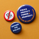 3 round pinback buttons, two small, one large. Defund the Police! in blue, Cop City with a red line slash through it & Police Protect Power & Property instead of our comities in white & gold on dark blue, the largest button.