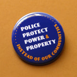 A round navy blue pinback button that reads Police Protect Power & Property, Instead of Our Communities in white & golden yellow text. Button is on a warm sand oat colored background.