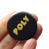 Round black pinback button that reads POLY in yellow, blue & pink overlapping san serif fonts. Pinback button is held in a hand.