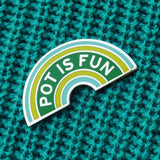 Hard enamel pin in the shape of a rainbow that reads POT IS FUN in silver type, with pale cyan, neon yellow and green enamels making up the layers of the rainbow, separated by thin silver outlines. Pinned to a teal blue green sweater.