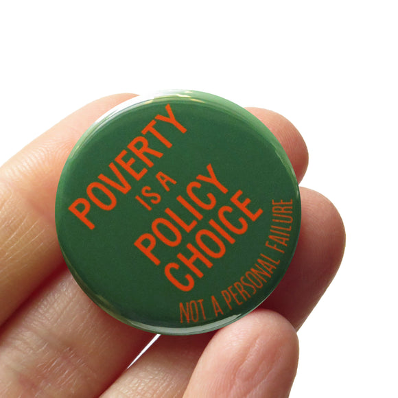 a round dark green pinback buton that reads POVERTY IS A POLICY CHOICE, Not a Personal Failure in orange text. Button is held in a hand.