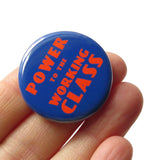 Round blue pinback button with red-orange text that reads POWER TO THE WORKING CLASS in an art deco font. Button is held in a hand.