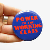 A round bold blue pinback button that reads POWER TO THE WORKING CLASS in red-orange text,  an art deco font. Button is held in a hand.