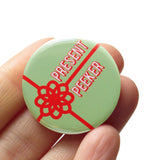 Round mint green pinback button with a bright red package bow and the words PRESENT PEEKER in white with a red outline text. Button is held in a hand.