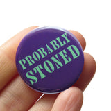 Round dark purple pinback button that reads PROBABLY STONED in minty green text. Badge is held in a hand