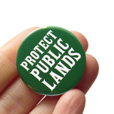 A round hunter green 1.25" pinback button that reads PROTECT PUBLIC LANDS in a white fonts. The pinback button is attached to a SOCIAL ALERT BUTTON backing card. 