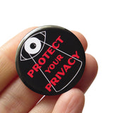 Round black pinback button that reads PROTECT YOUR PRIVACY in red text with a big brother overseer eye  overlooking the text. Button is held in a hand.