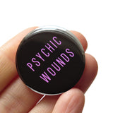 Round black 1.25" pinback button that reads PSYCHIC WOUNDS in a thin lavender font. Badge is held in a hand