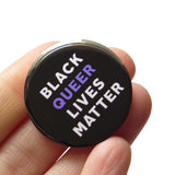 Round 1.25" black pinback button that reads BLACK QUEER LIVES MATTER in white & purple text. Badge is held in a hand.