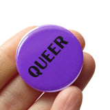 ound purple pinback button that reads QUEER in black text. Held in a hand.