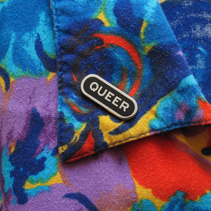 Capsule shaped silver pin with black enamel that reads QUEER. Pin is on the lapel of a colorful vintage shirt.