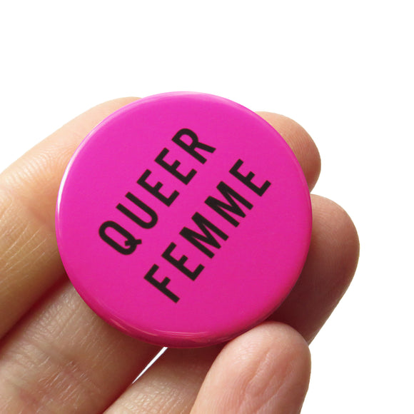 Magenta pink round pinback button that reads QUEER FEMME in black san serif text. Badge is held in a hand.