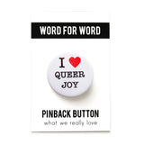 A round white pinback button that reads I LOVE QUEER JOY in a black serif font, the love being indicated by a red heart. 