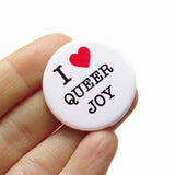 A round white pinback button that reads I LOVE QUEER JOY in a black serif font, the love being indicated by a red heart.  Button is held in a hand.