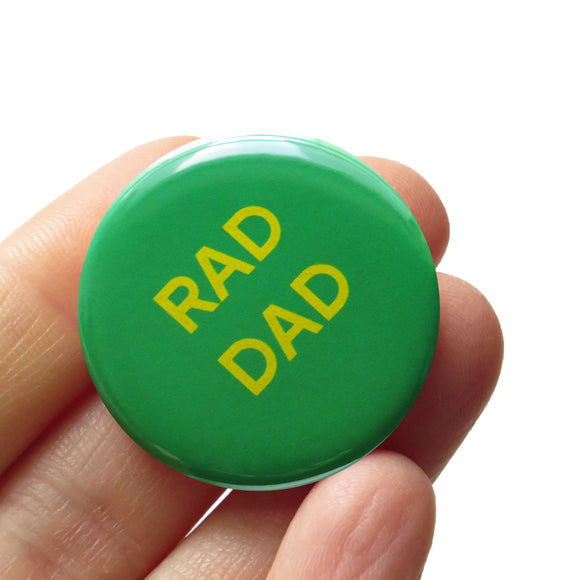 Round Keely green pinback button with yellow text that reads RAD DAD. Badge is held in a hand.