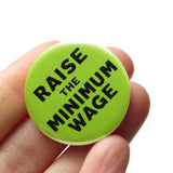 Round green pinback button that reads RAISE THE MINIMUM WAGE in black text. Button is held in a hand.