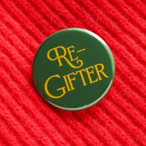 Rich green round pinback button that reads RE-GIFTER in decorative golden yellow font. Button is on a bright red soft sweater with a diagonal rib pattern.