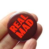 A dark brown pinback button with thick lava orange text that reads REAL MAD in two lines taking up all the space on the face of the button. Button badge is held in a hand.