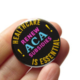 a round black pinback button with yellow, magenta and cyan text that reads HEALTHCARE IS ESSENTIAL, Renew ACA Subsidies. Button is held in a hand.