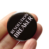 Shiny round black 1.25" pinback button, which reads RESOLUTION BREAKER in white text. Button is held in a hand.