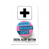 A round pink pinback button with light aqua text and dark teal boxes that reads REVERSE MEDICAID CUTS. Button is on a social alert button backing card.